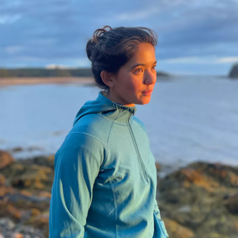 The image shows a woman standing near the ocean. She is wearing a blue jacket and looking off to the side. The background includes the ocean, rocks, and a cloudy sky. The lighting suggests it might be sunrise or sunset.